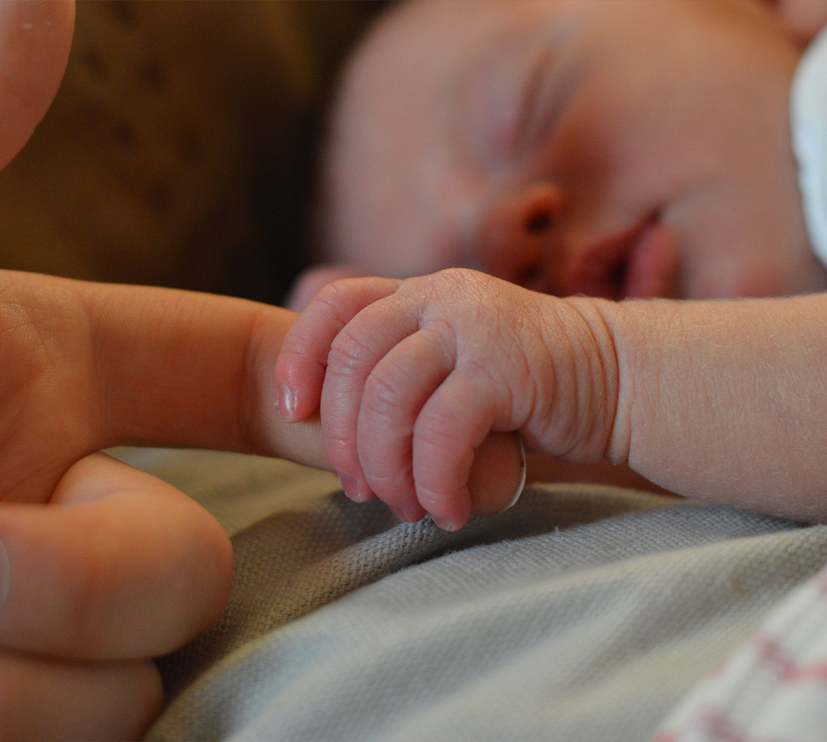 A tiny newborn grasps her caregiver's index finger while sleeping
